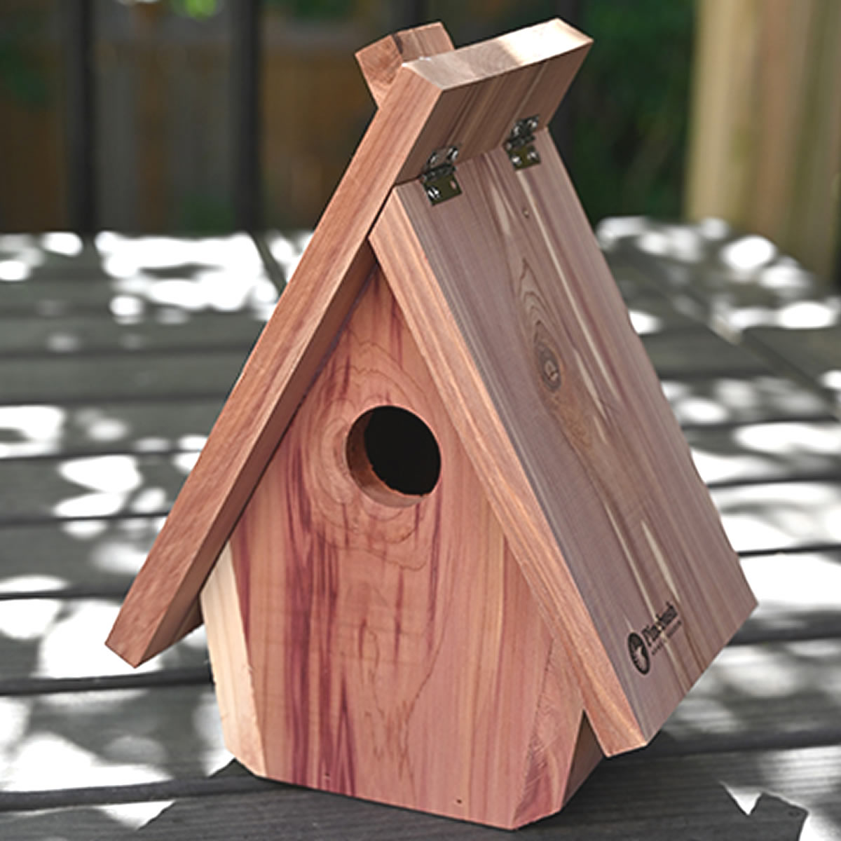 Chalet Style Nesting Box featuring a simple A-frame roof with metal hinges, a round entrance hole, on a wood-plank surface in dappled sunlight.