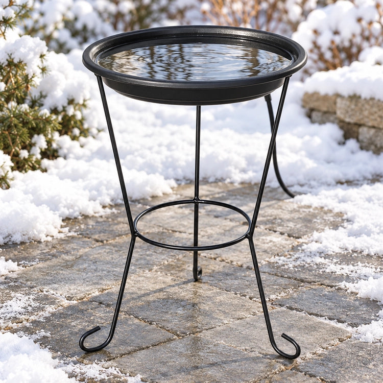 Black heated bird bath with clear water on a three-legged metal stand, surrounded by snow on a stone patio