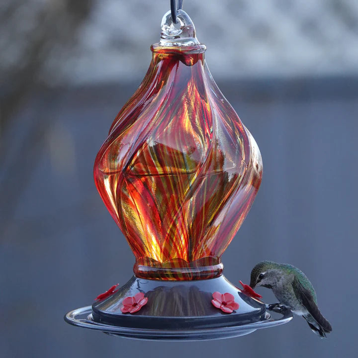 EcoCycle Sunburst Sipper handblown recycled glass hummingbird feeder with swirling amber, orange, and red glass design, visited by a hummingbird at a flower-shaped feeding port.