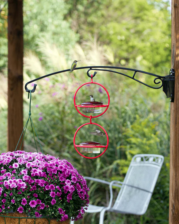 Brome BirdsUP 25 Hanging Bracket holds two hummingbird feeders and a flower basket, showcasing its sturdy design amidst a garden patio scene.
