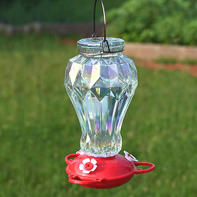 Iridescent Glass Hummingbird Feeder with faceted glass reservoir, metal hanger, and red base with white flower ports, hanging above a lawn.