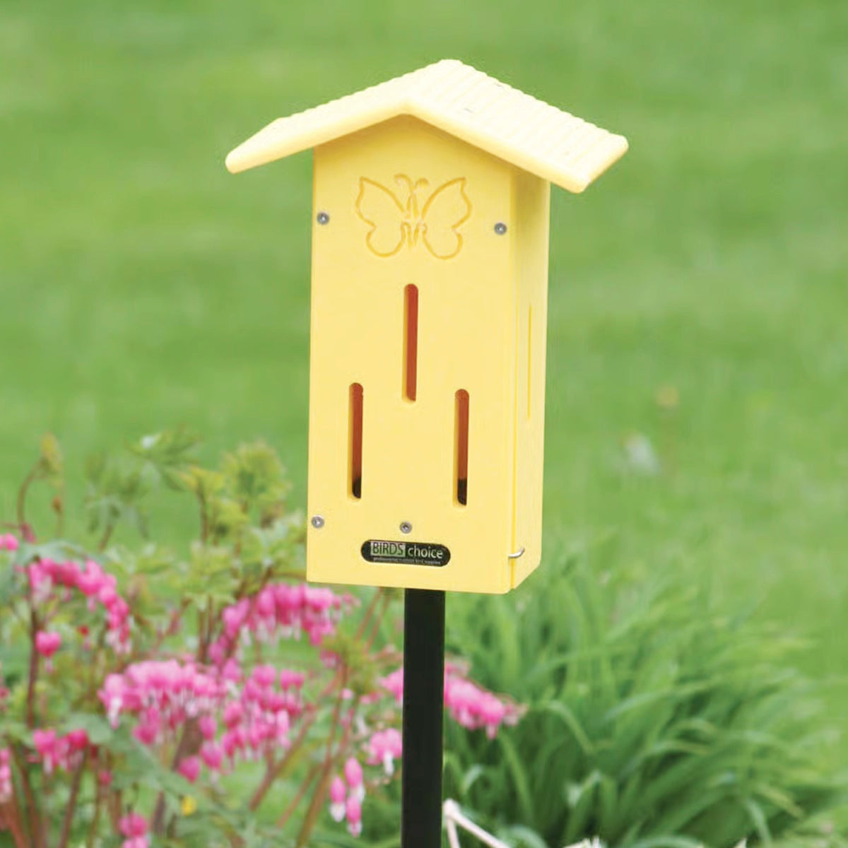 BirdsChoice Pole-Mounted Butterfly House, a yellow insect shelter on a black stake, among pink flowers in a garden, features decorative butterfly design.