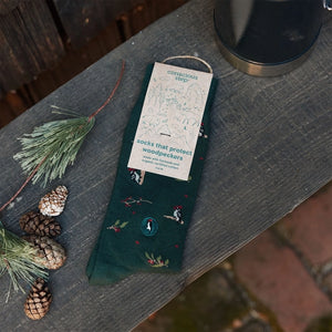 Conscious Step woodpecker socks with eco-friendly packaging displayed alongside pine branches and pinecones