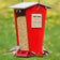 Snack Shack Resistor Feeder: Red, house-shaped bird feeder with clear panels, two perches, filled with seed; titmouse and goldfinch visible, squirrel-resistant design.
