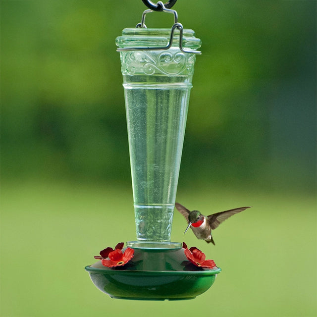 8 oz. Torchiere Glass Hummingbird Feeder with red flower ports and embossed green glass, attracting a hummingbird as it hovers to drink nectar.