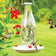 Milkhouse 24 Ounce Glass Hummingbird Feeder, featuring a vintage milk can design, with red flower feeding ports and hummingbirds feeding, against a green backdrop.