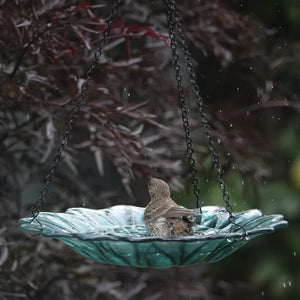 Turquoise recycled glass hanging bird bath displayed outdoors with decorative stones in the basin, surrounded by garden foliage.