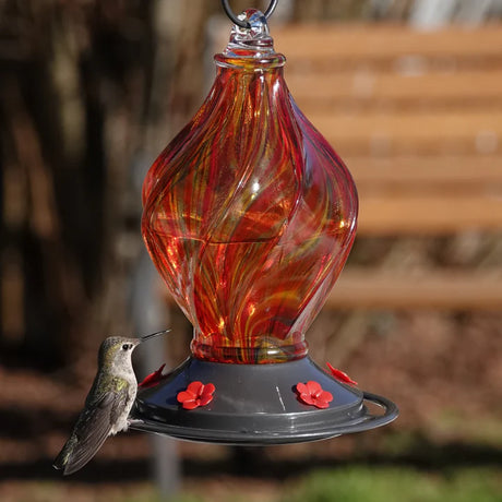 EcoCycle Sunburst Sipper handblown recycled glass hummingbird feeder with swirling amber, orange, and red glass design, visited by a hummingbird at a flower-shaped feeding port.