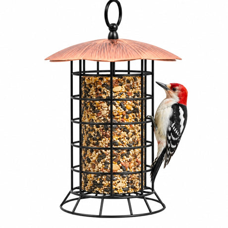 A cylindrical bird feeder with a copper-colored metal roof and a black wire cage body, shown on a white background with a red, black, and white woodpecker perched on the right side.