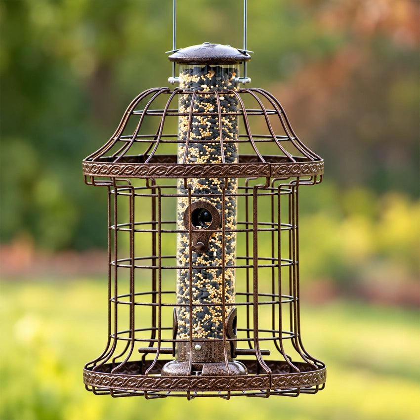 Decorative bronze squirrel-resistant bird feeder with protective metal cage and seed-filled tube, hanging in a beautiful green backyard setting.