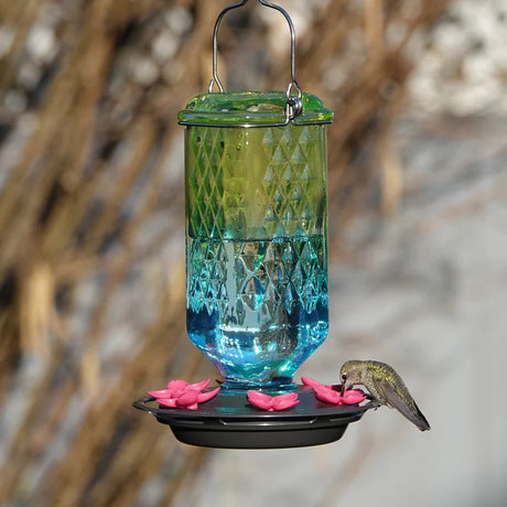 Seafoam Ombre Column Antique Hummingbird Feeder hanging outdoors, featuring an embossed glass reservoir with a seafoam green to blue ombre finish and flower-shaped feeding ports.