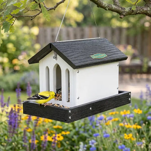 Bird feeder hanging from a tree with a bird perched on it, surrounded by flowers and greenery.