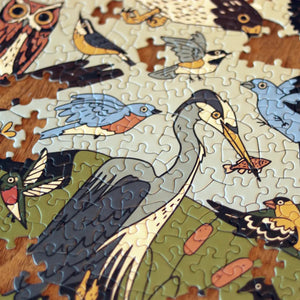 Partially assembled Birds of Eastern North America Puzzle, showcasing various stylized birds, like herons and songbirds, against a muted background on a wooden surface.