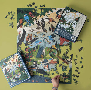 Birds of Eastern North America Puzzle partially assembled, showing diverse bird species. Puzzle box nearby, hand placing final piece, magnifying glass rests on corner.